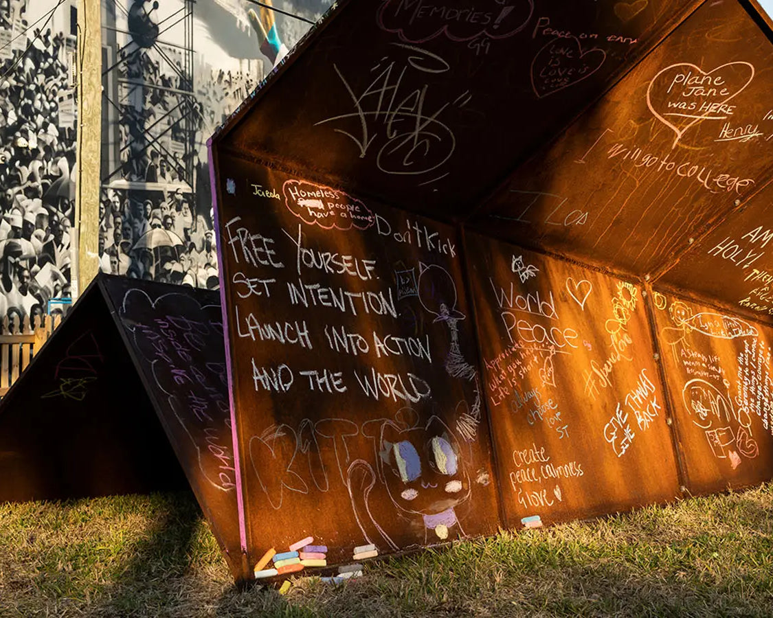 Rust-colored metal paper airplane sculpture with local children's intention's written on it in chalk on a grassy area for Canvas Outdoor Museum in Palm Beach, FL.