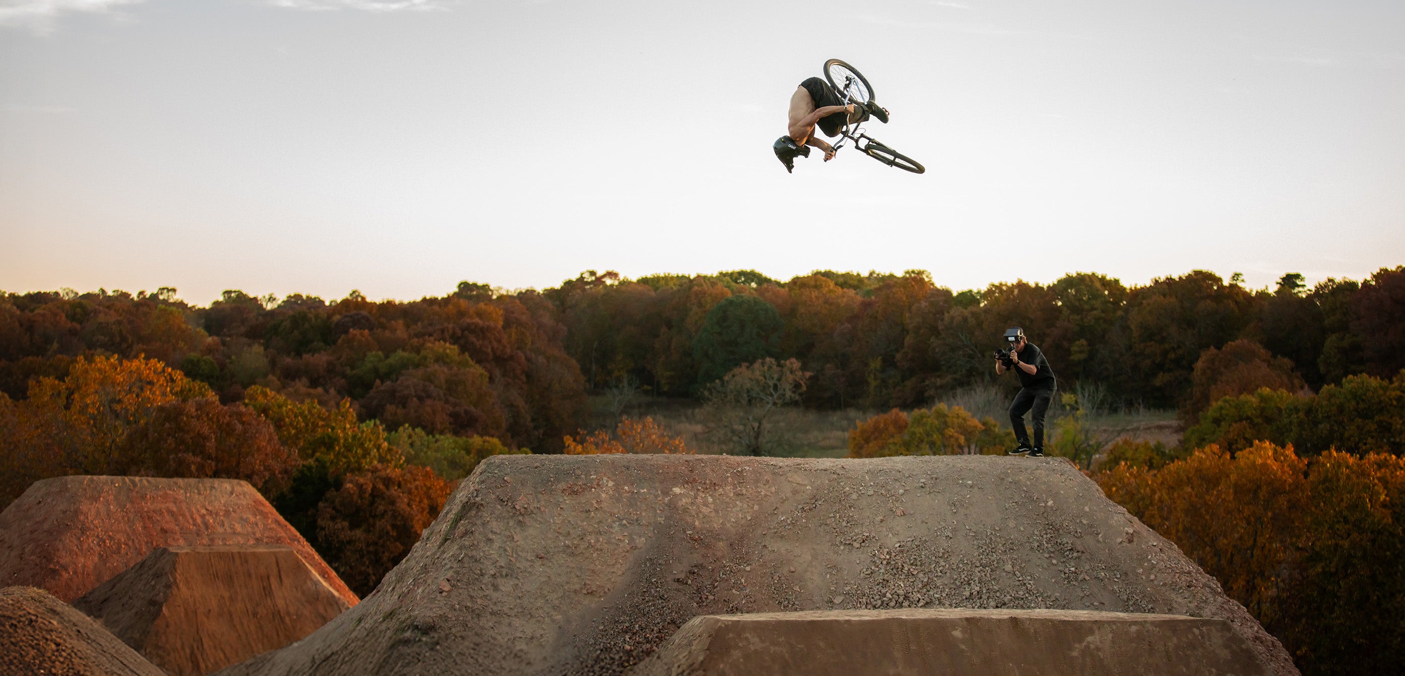 Person on a mountain bike performing a jump over a dirt ramp with trees in the background at the Drill course built by Launch Intention's Griffin Loop.