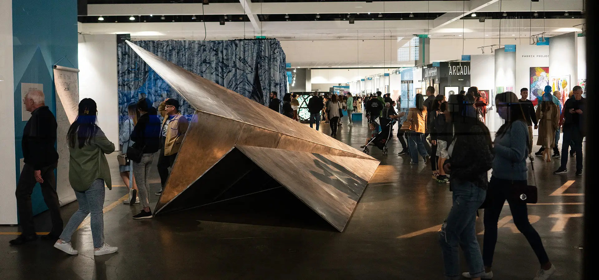 Large metallic paper airplane sculpture in a modern indoor setting with people around at the LA Art Show.