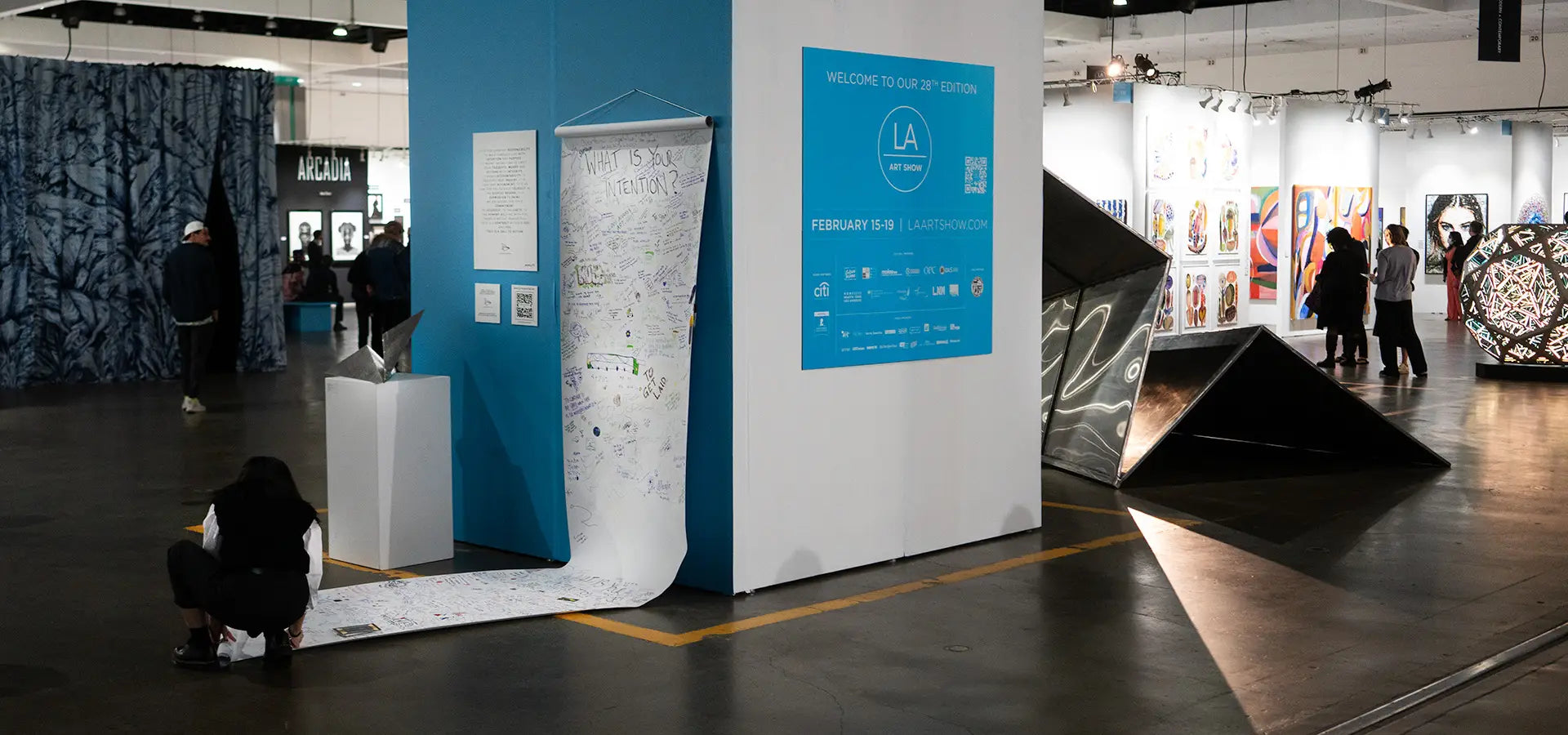 Large Launch Intention metallic paper airplane sculpture in a modern indoor setting at the LA Art Show as show goers write their intentions on a scroll that extends down a wall onto the floor.