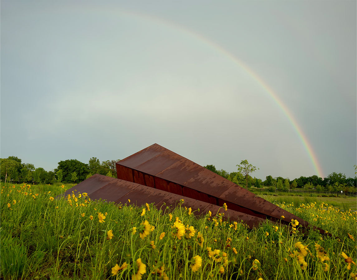 25-foot Launch Intention paper airplane sculpture by Griffin Loop in Osage Park framed by wildflowers and a rainbow, public art installation inspiring intention and community connection 