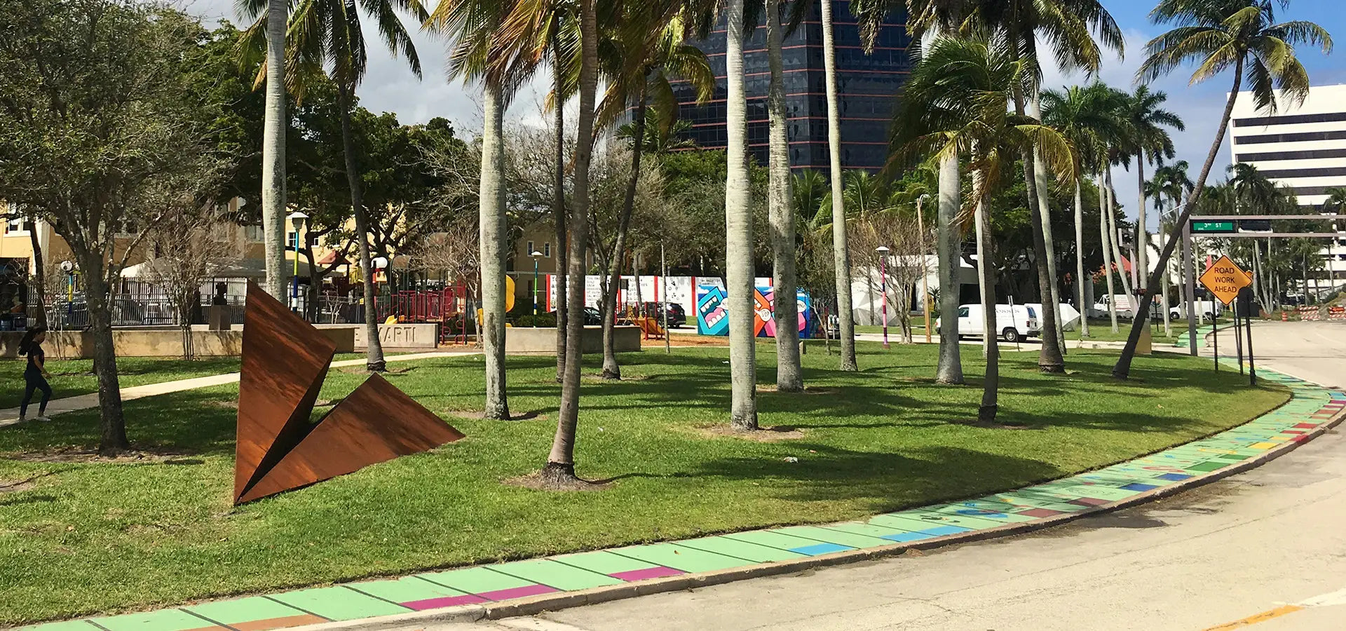A Metal Paper Airplane sculpture in a Florida Park with palm trees, a colorful pathway during the Canvas Outdoor Museum Festival.