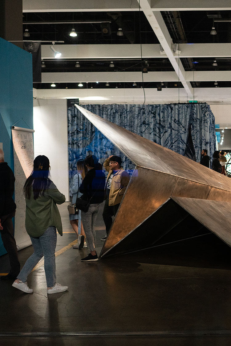 Large metallic paper airplane sculpture in a modern indoor setting with people around at the LA Art Show.