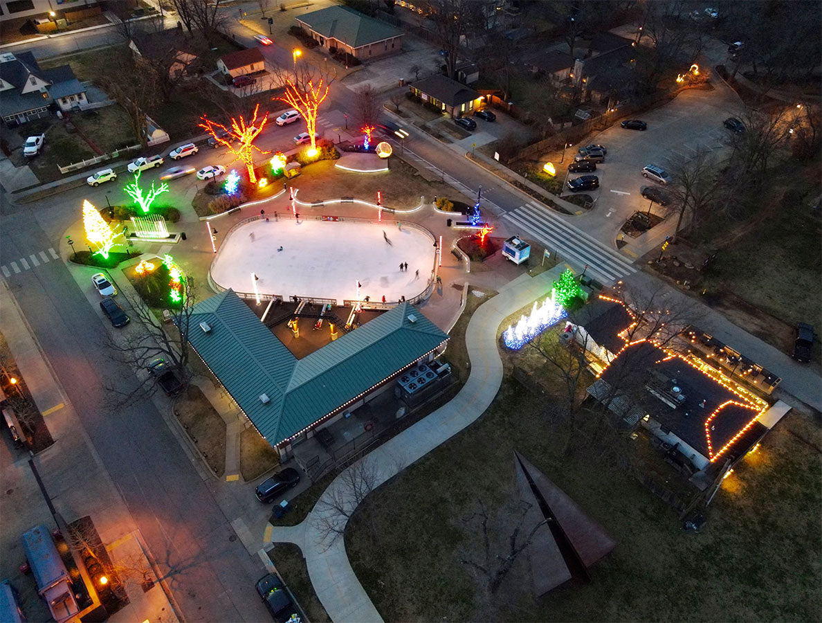  50-foot Launch Intention paper airplane sculpture by Griffin Loop in downtown Bentonville at night, carnival lights and ice rink activity creating a vibrant public art gathering space 