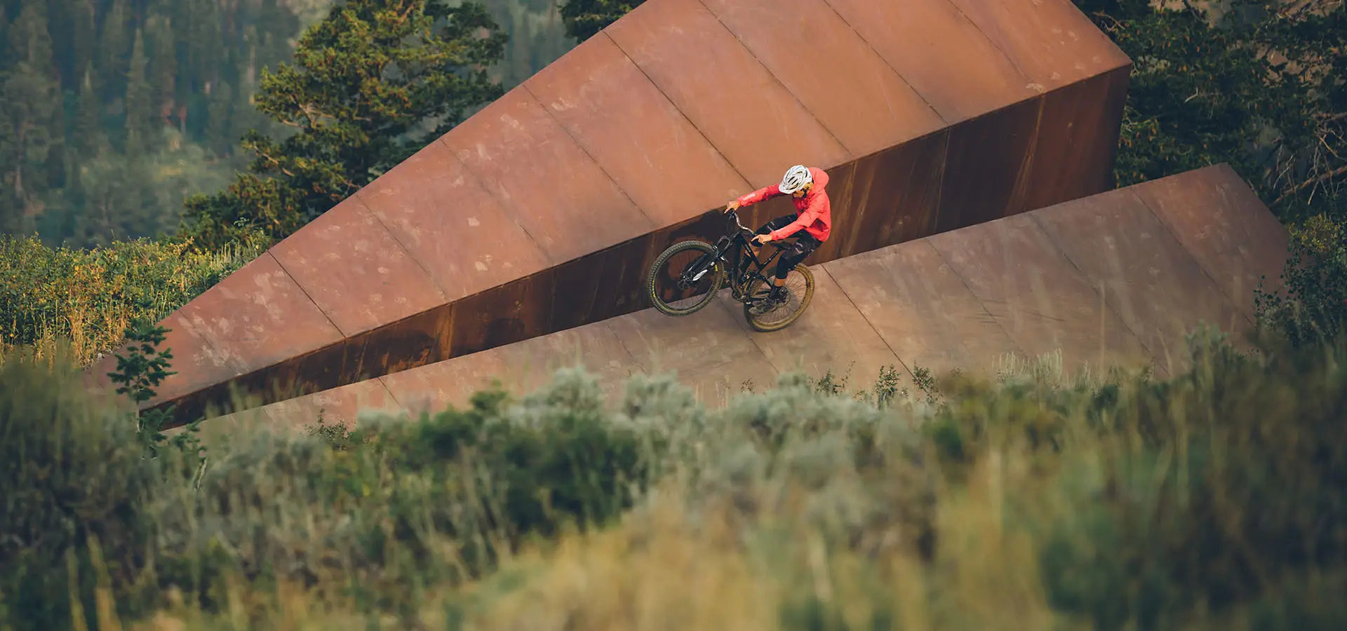 Person on a mountain bike riding across Powder Mountain's 50ft Launch Intention metal paper airplane sculpture on top of the mountain.