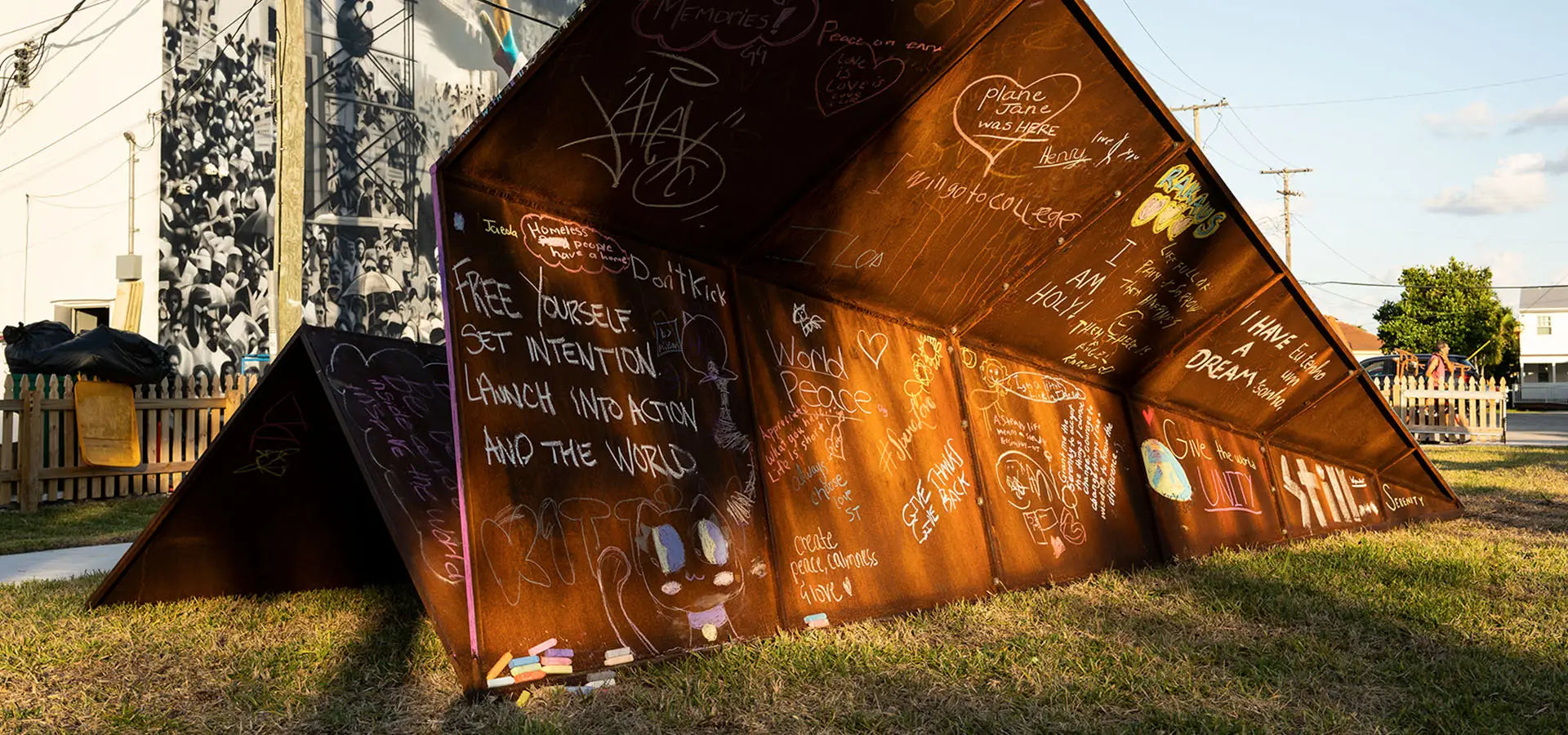 Rust-colored metal paper airplane sculpture with local children's intention's written on it in chalk on a grassy area for Canvas Outdoor Museum in Palm Beach, FL.