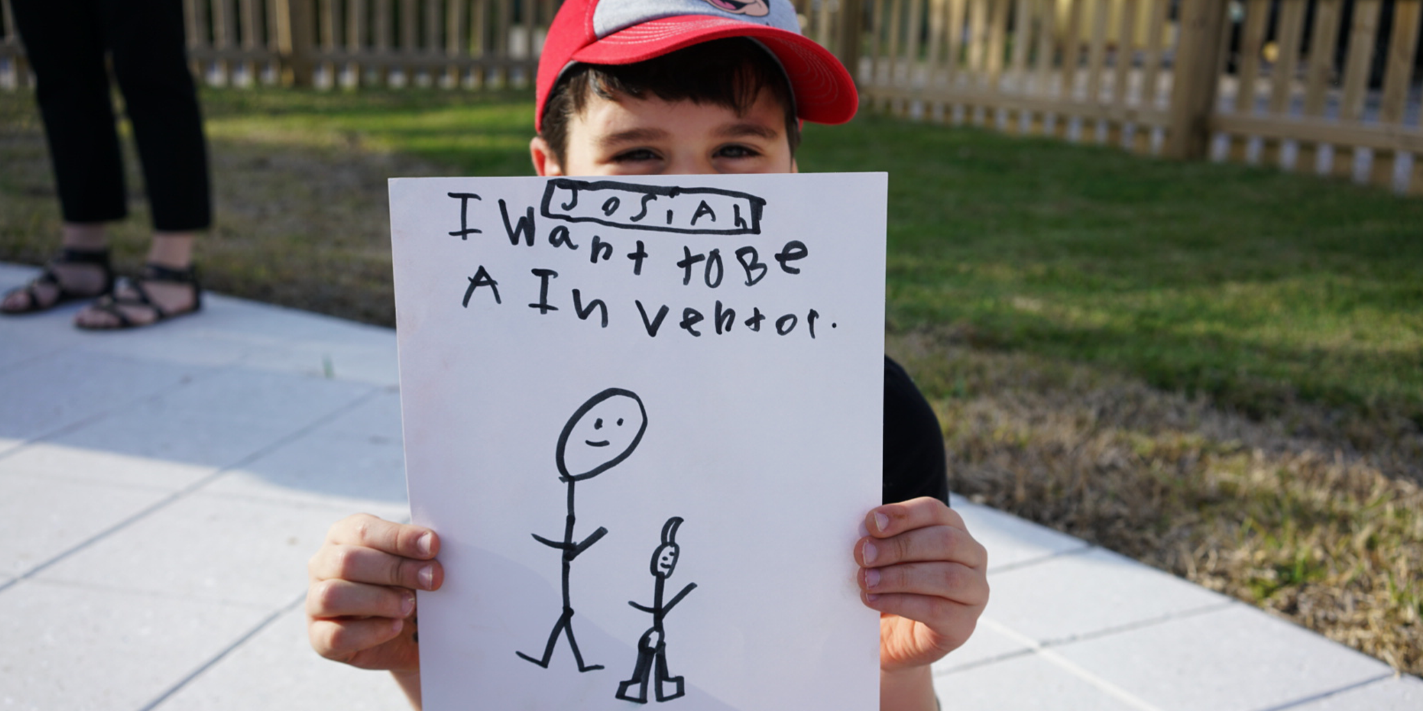 Child holding a paper with a drawing of a stick figure and text of his intentions outdoors.