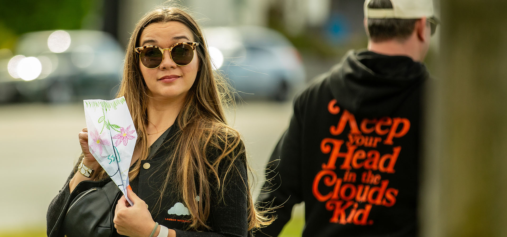 Woman holding a colorful paper airplane with a blurred background of a person wearing a hoodie with text reading "Keep Your Head in the Clouds Kid." from Launch Intention at the San Francisco Marina Green sculpture instalation.
