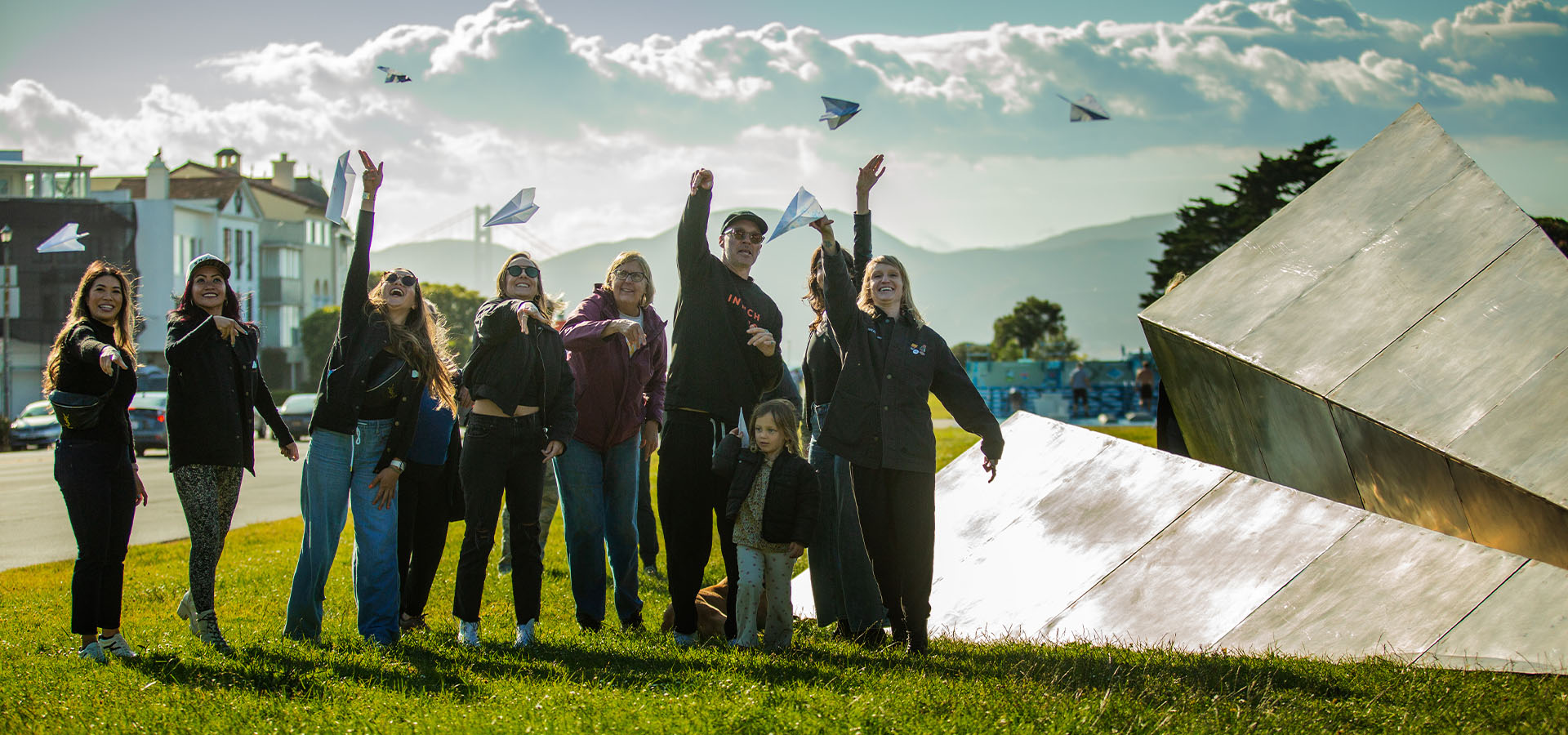 Group of people throwing Launch Intention paper airplanes next to the Launch Intention 25 Ft Paper Airplane sculpture on the Marina Green Triangle and the Golden Gate Bridge in the background.