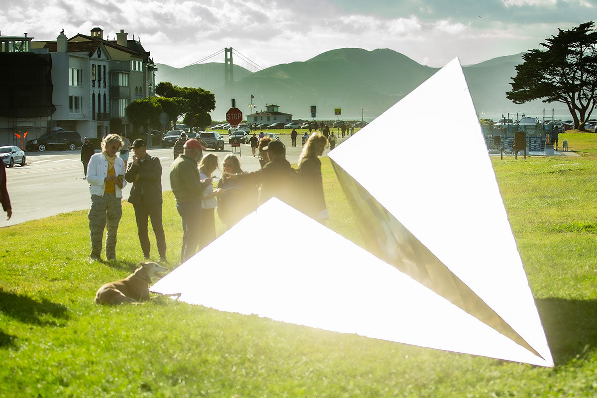 People and a dog celebrating the Launch Intention 25 Ft Paper Airplane sculpture beaming in sunlight on the Marina Green Triangle opening with the Golden Gate Bridge in the background.
