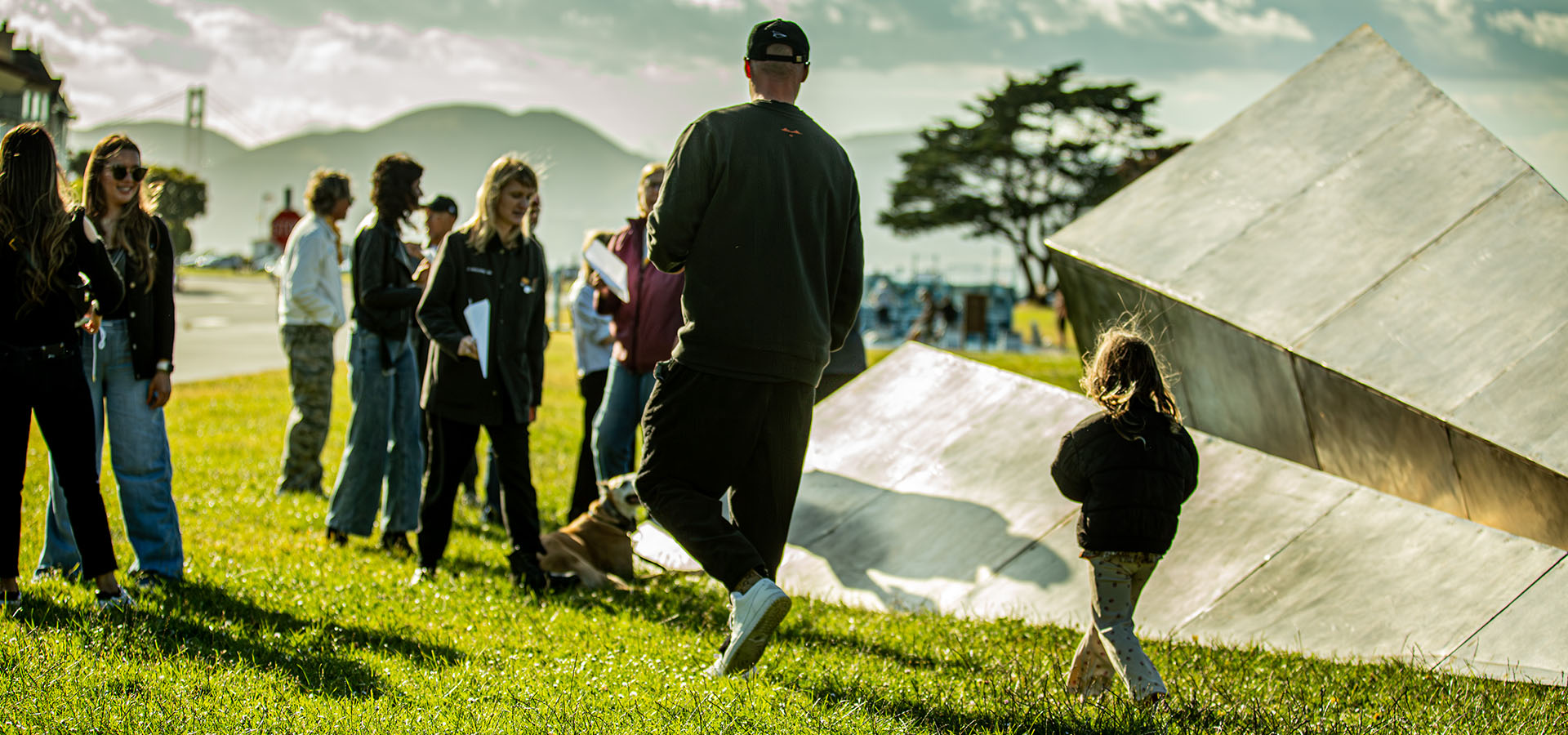 Group of people standing on grass near the Launch Intention 25 Ft Paper Airplane sculpture beaming in sunlight on the Marina Green Triangle opening with the Golden Gate Bridge in the background.